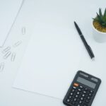 A desk scene with blank papers, a calculator, a pen, paper clips, and a small plant.