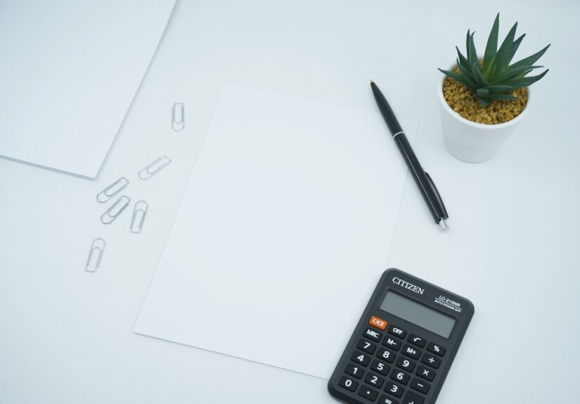 A desk scene with blank papers, a calculator, a pen, paper clips, and a small plant.